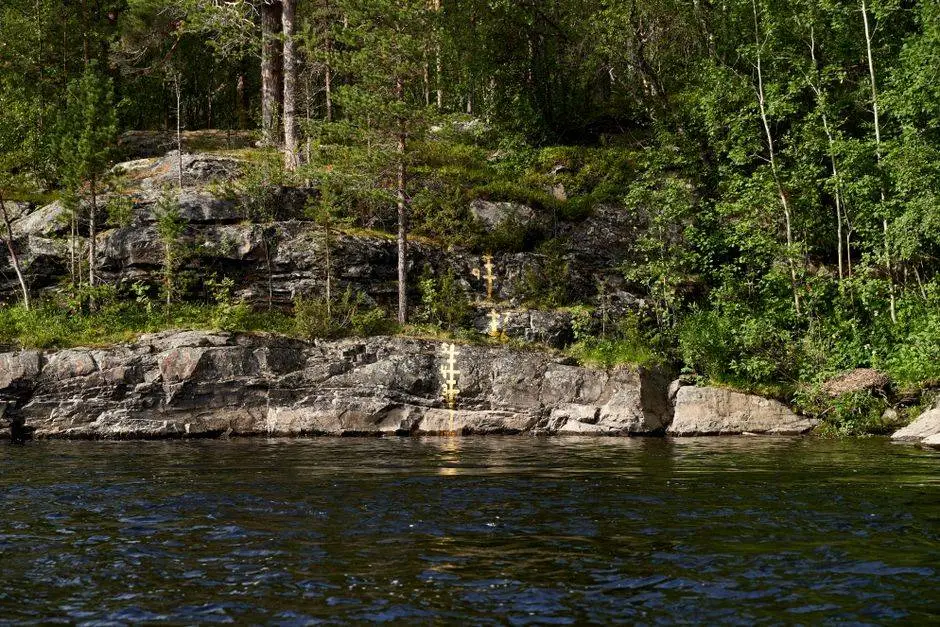 Peaceful lakeshore scene featuring rocky terrain and lush green forest under a clear blue sky.